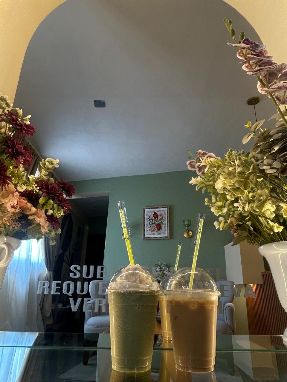 Bebidas de matcha y café helado sobre una mesa de vidrio, rodeadas de flores.